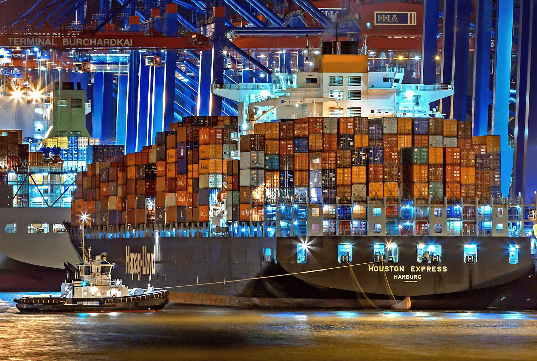 A large cargo ship loaded with containers is docked at a brightly lit port at night, while a small tugboat is positioned near the ships hull. Cranes and terminal signs are visible in the background.