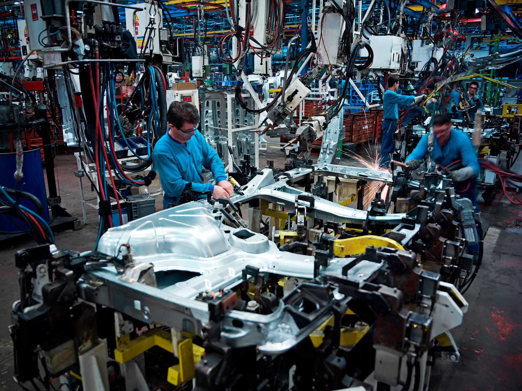 Workers in blue uniforms assemble car frames on a busy automotive factory floor, surrounded by machinery, tools, and robotic arms, with sparks flying from welding in the background.