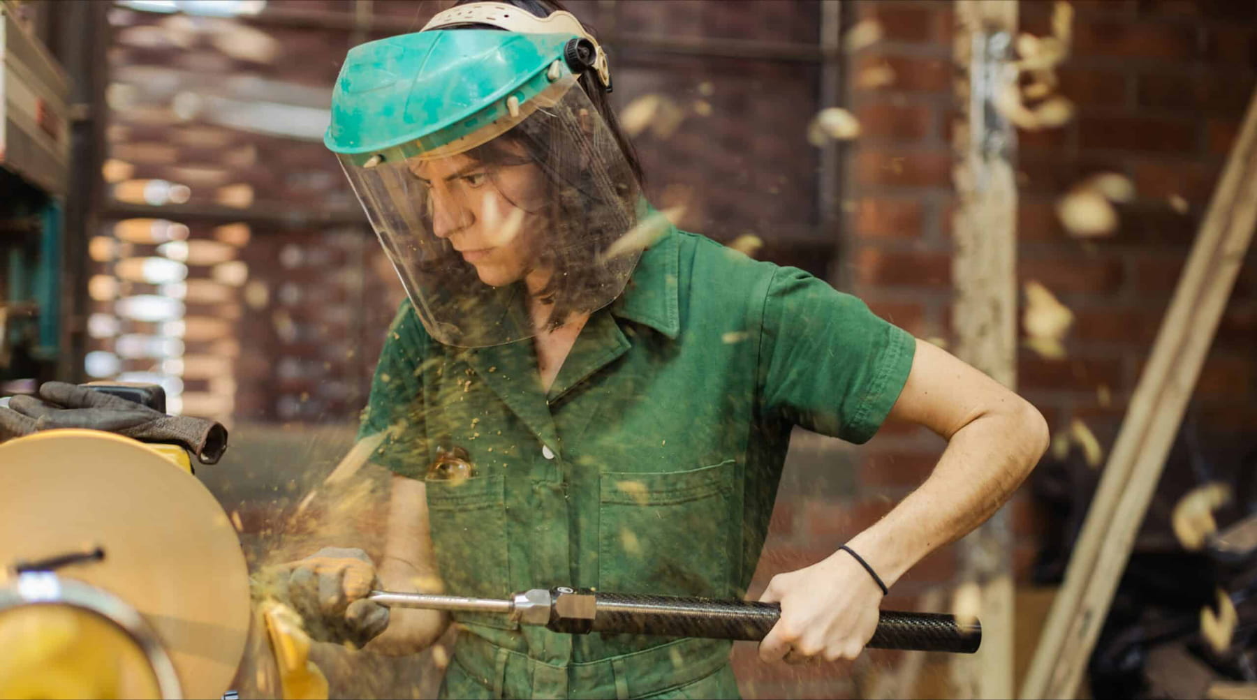A person wearing a green jumpsuit and protective face shield operates a lathe, shaping a piece of wood as wood chips fly through the air in a workshop with brick walls.