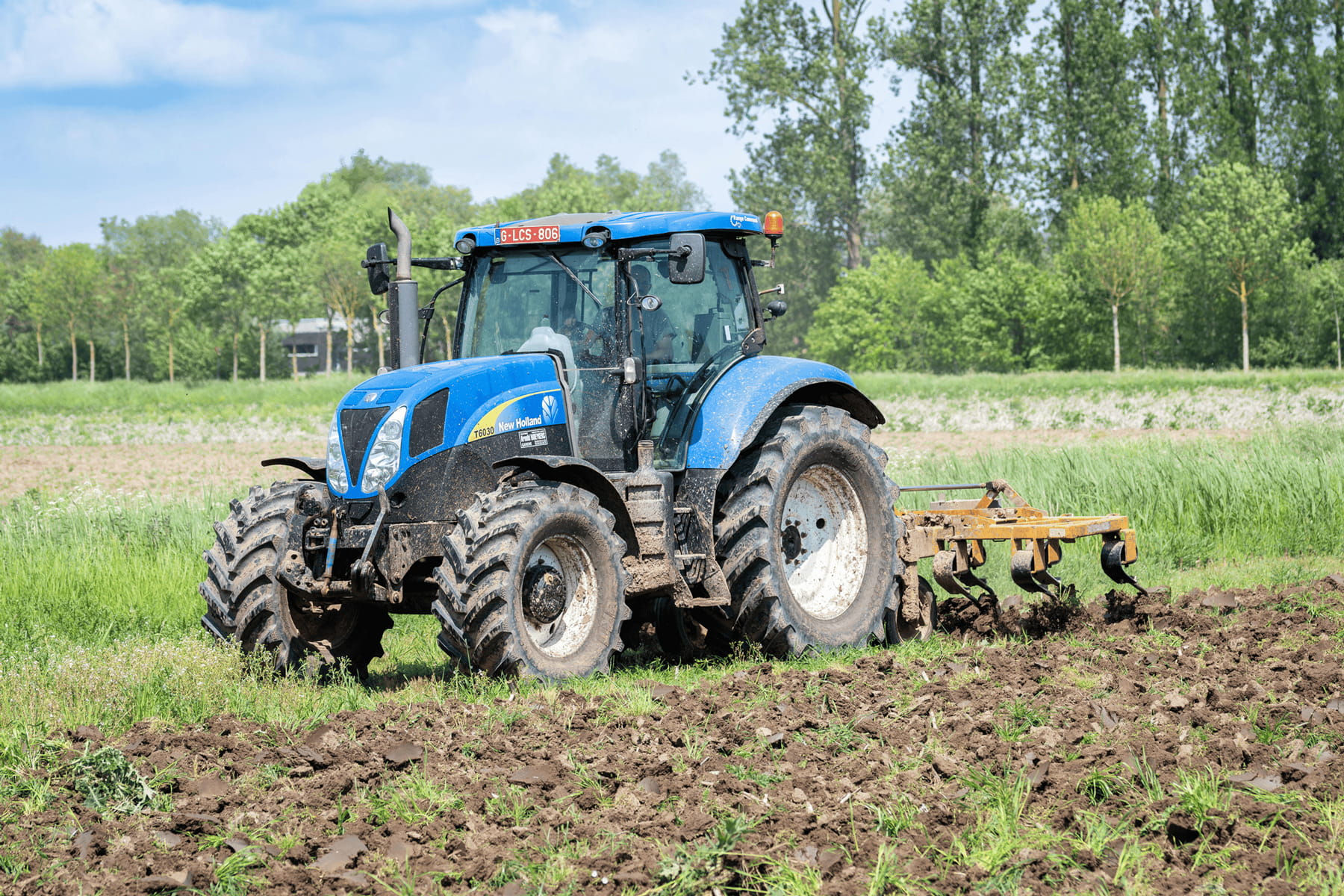 A blue tractor plows a muddy field on a farm, surrounded by green grass and trees under a partly cloudy sky.