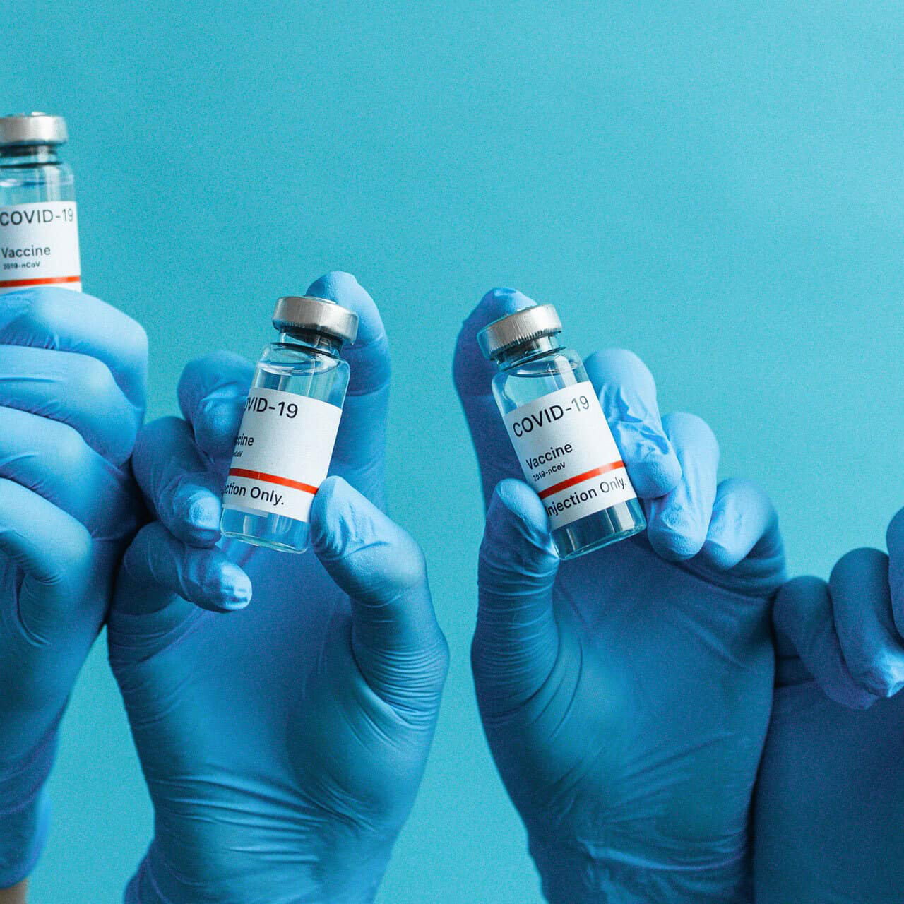 Four hands wearing blue medical gloves hold up vials labeled COVID-19 Vaccine against a solid blue background, symbolizing vaccination and healthcare efforts against the coronavirus.