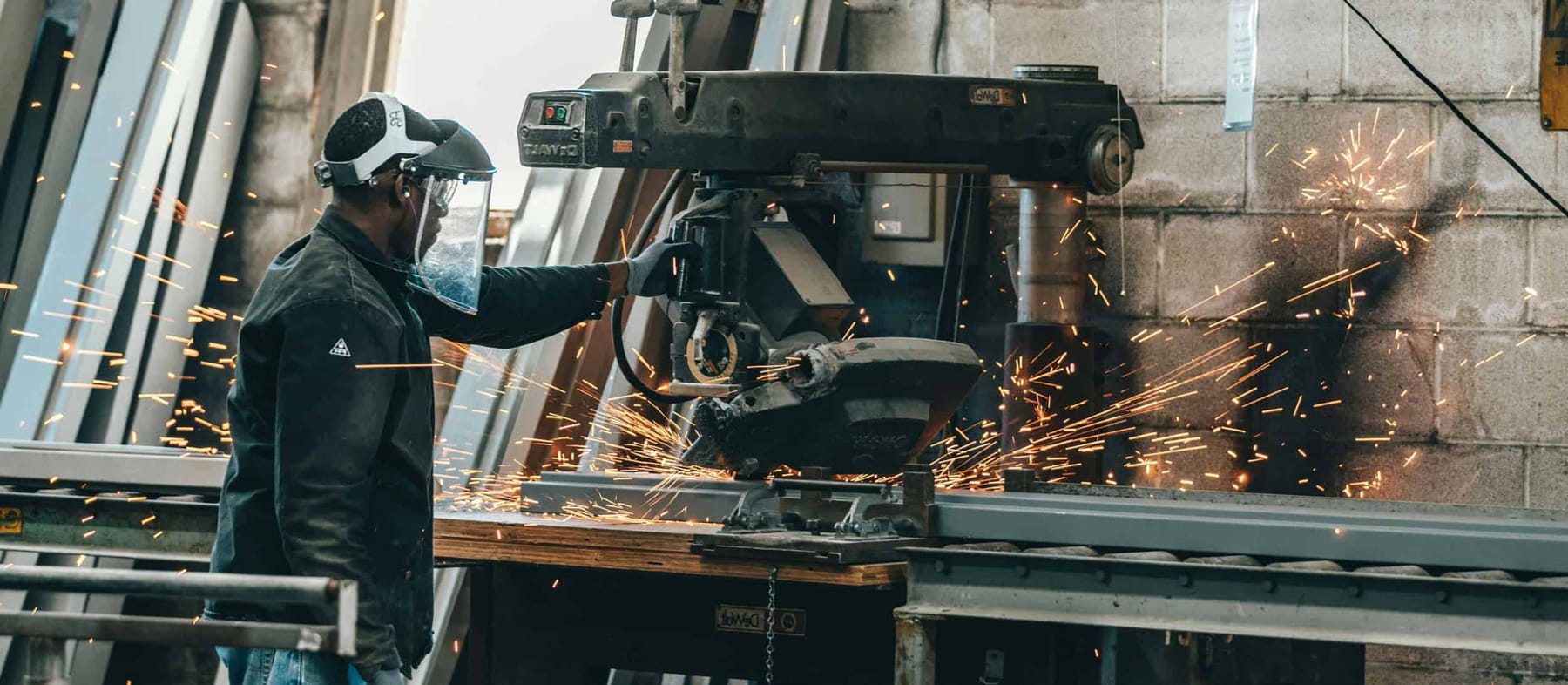 A worker wearing protective gear operates a large metal cutting machine, causing bright sparks to fly, in an industrial workshop with metal beams and concrete walls.
