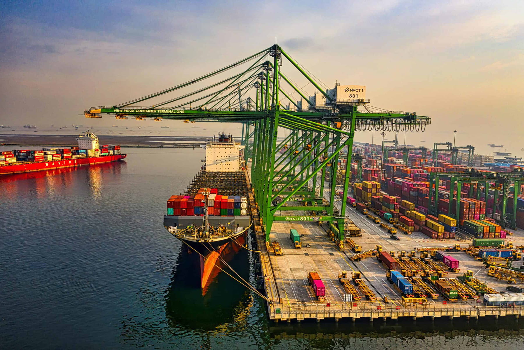 A large container ship is docked at a busy port, surrounded by cranes and stacks of colorful shipping containers. Another ship sails nearby, with more containers visible in the background under a hazy sky.