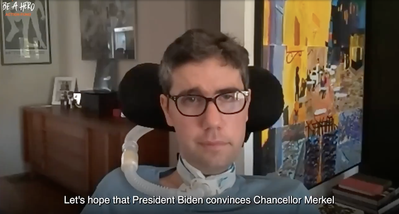 A man with glasses and a ventilator sits indoors, with colorful artwork and framed photos behind him. Subtitles read: Lets hope that President Biden convinces Chancellor Merkel.