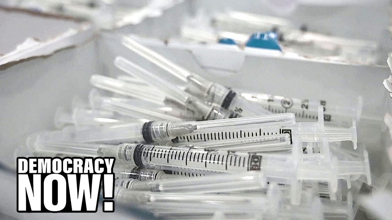 A pile of unused medical syringes is shown in a box. The words DEMOCRACY NOW! are displayed in bold white text in the bottom left corner of the image.