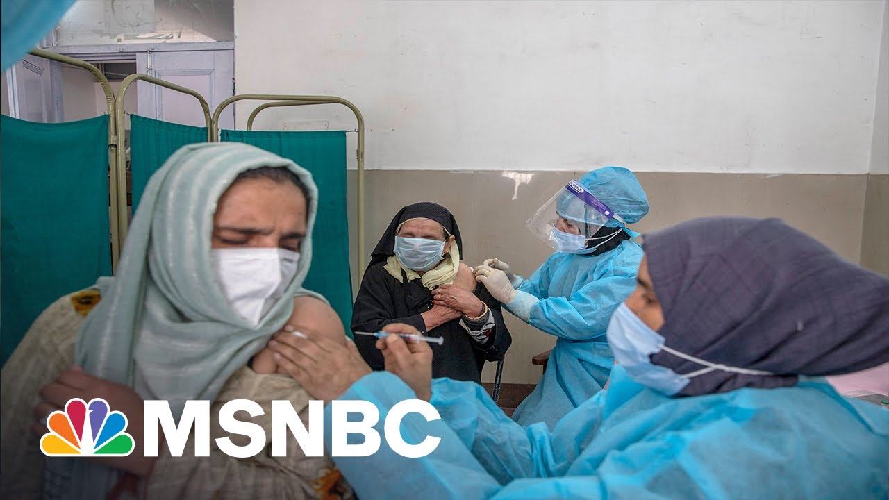 Healthcare workers in blue protective gear administer vaccines to women in headscarves in a clinical setting. The MSNBC logo is overlaid in the bottom left corner.